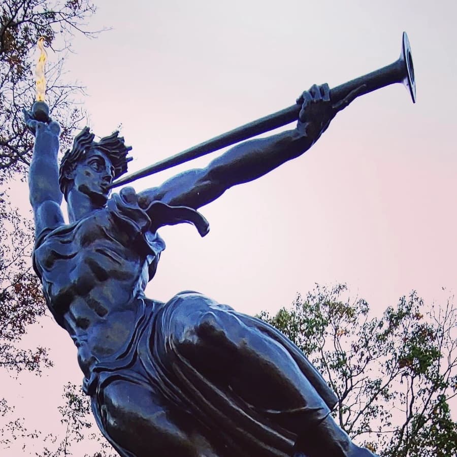 A bronze statue of a woman holding a torch and a trumpet, poised in a dynamic pose against a pastel sky.